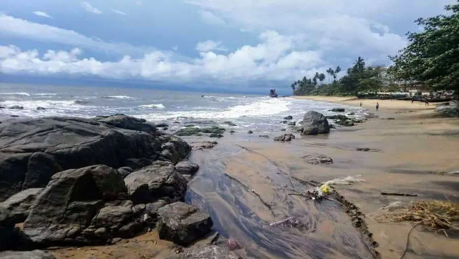 Plage d'Ebodjé, rochers noirs et vagues sous un ciel nuageux